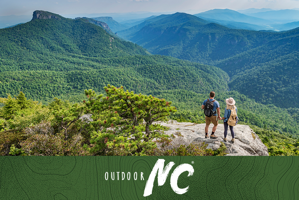 Aerial view of couple standing on an overlook at Hawksbill Mountain in daytime along the Blue Ridge Mountain range.