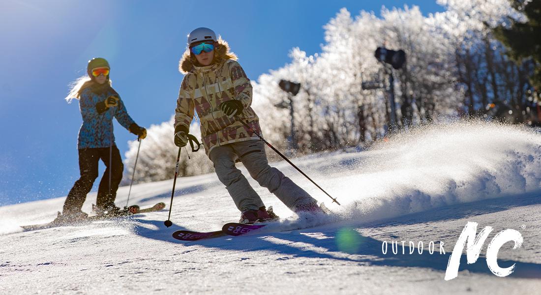 OUTDOOR NC - Two people ski down a snowy slope on a sunny day