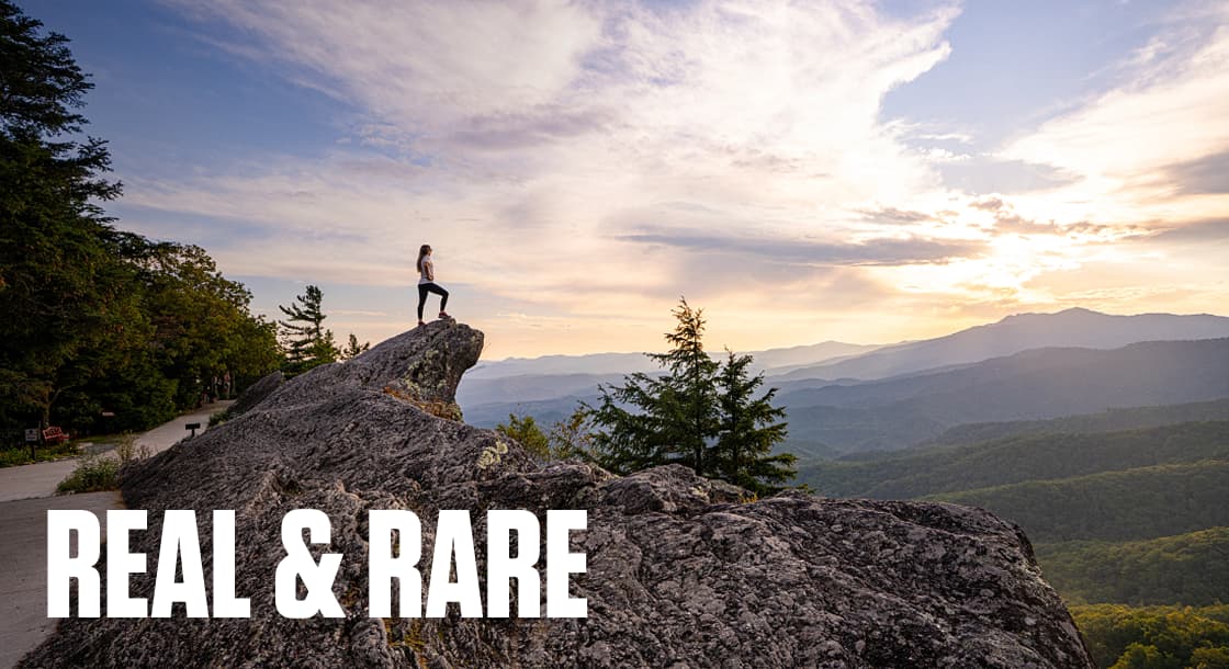 REAL & RARE - Scene showing a young woman standing on The Blowing Rock at dusk with mountains in the background. Blowing Rock, NC