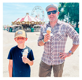 Adult man and young boy smiling and holding ice cream cones in front of a carnival