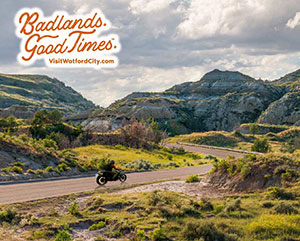 Wide view of person riding a motorcycle through rocky and grassy landscapes, with overlay text reading: Badlands. Good Times. VisitWatfordCity.com