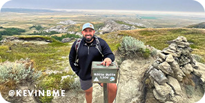 Smiling man standing on top of a mountain with green and rocky landscapes behind him and text overlay that reads @Kevinbme