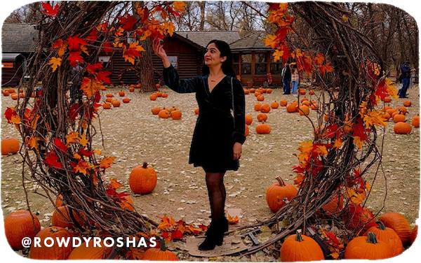 A woman wearing a black dress standing in a large wreath of fall foliage and surrounded by pumpkins, with overlay text that reads @rowdyroshas