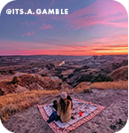 A woman sitting on a blanket overlooking a beautiful pink sunset, with overlay text that reads @its.a.gamble