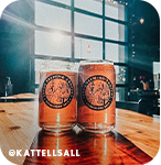 Two orange beer cans sitting on a table in front of a glass garage door at a brewery, with overlay text that reads @kattellsall