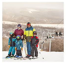 Smiling family of five people standing on skis wearing ski gear in front of snowy terrain and a ski lift