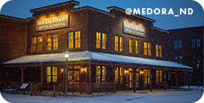 Brick building at night with illuminated windows on a snowy street, with overlay text that reads @medora_nd