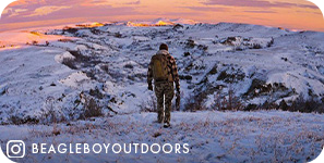 View from behind of a man trekking through snow with snowy mountains and a pink sunset ahead of him, with overlay text that reads @beagleboyoutdoors