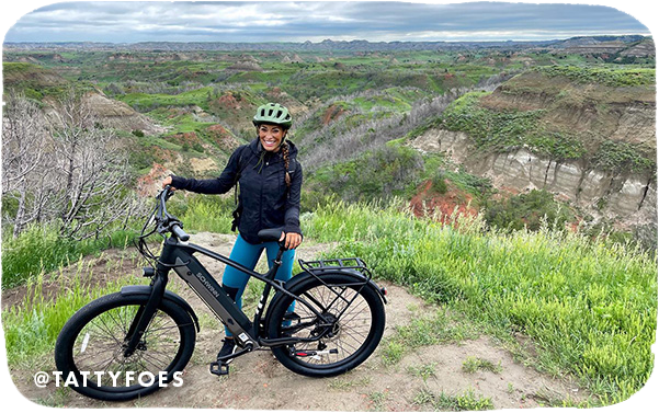 Woman posing with a mountain bike at a vista point overlooking green hills, with overlay text that reads: @tattyfoes