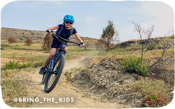 Young child riding a mountain bike on a dirt trail, with overlay text that reads: @bring_the_kids