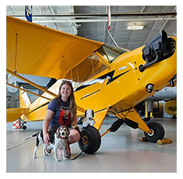 Woman kneeling next to a small yellow airplane with a dog next to her