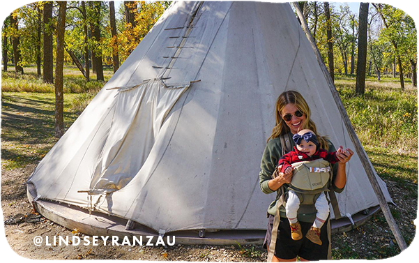 Woman holding a baby in a carrier while posing in front of a tent, with overlay text that reads: @lindseyranzau