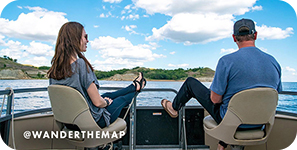 View from behind of a man and a woman sitting on a boat with water and blue sky ahead of them, with overlay text that reads: @wanderthemap