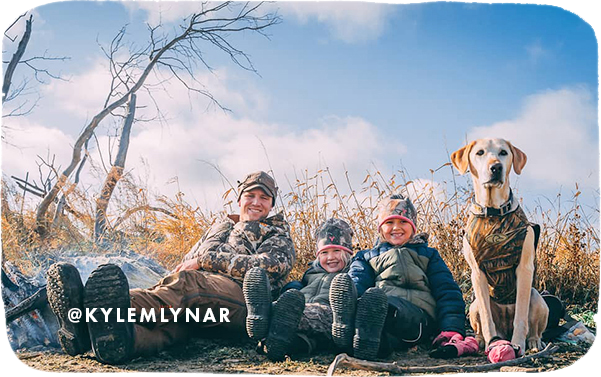 Man, two young girls and a dog sitting on the ground in a field wearing jackets and camouflage, with overlay text that reads: @kylemlynar