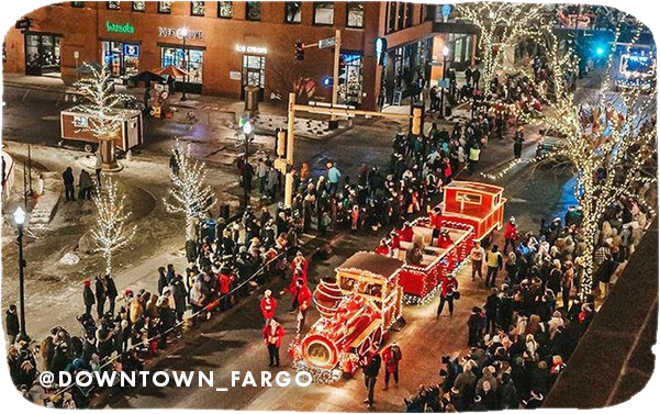 Aerial view of a holiday parade with holiday lights on a float, and overlay text that reads: @downtown_fargo
