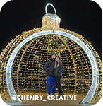 A man and a woman posing at night in a lit-up sculpture shaped like a round Christmas ornament, with overlay text that reads: @chenry_creative