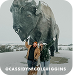 Two people and a dog posing in front of a large buffalo sculpture on a snowy day, with overlay text that reads: @cassidynicolehiggins