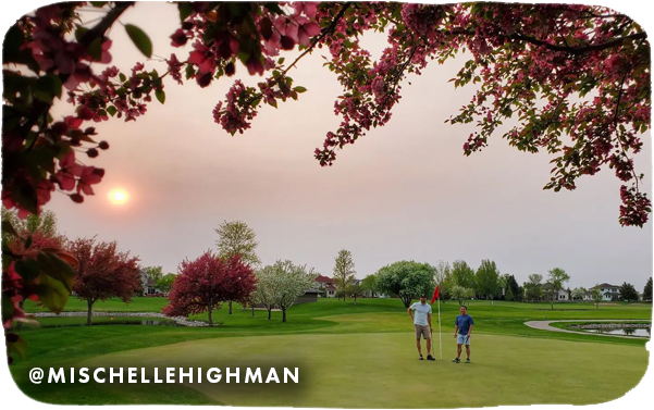 Two men standing on a golf green framed by tree branches with pink blossoms, with overlay text that reads: @mischellehighman