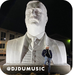 Man posing in front of a large, illuminated statue of Teddy Roosevelt at night, with overlay text that reads: @djdumusic