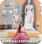 Woman in a pink dress posing in front of a mural of two women, with overlay text that reads: @sprinkledbymagic