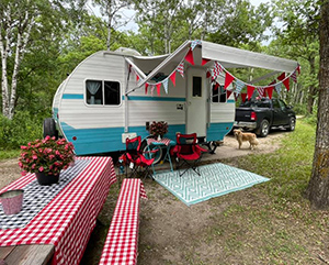 A white and teal camper trailer set up with an awning, camp chairs and a picnic table with a red tablecloth
