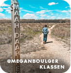 View from behind of a person walking on a trail in a barren landscape with a trail marker in the foreground that reads 