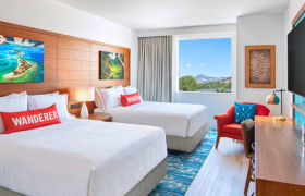 A bright, modern hotel room featuring two double beds with white bedding and red pillows.