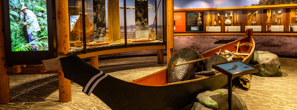 A museum exhibit showcasing a traditional wooden canoe with informational displays and a video screen in the background.