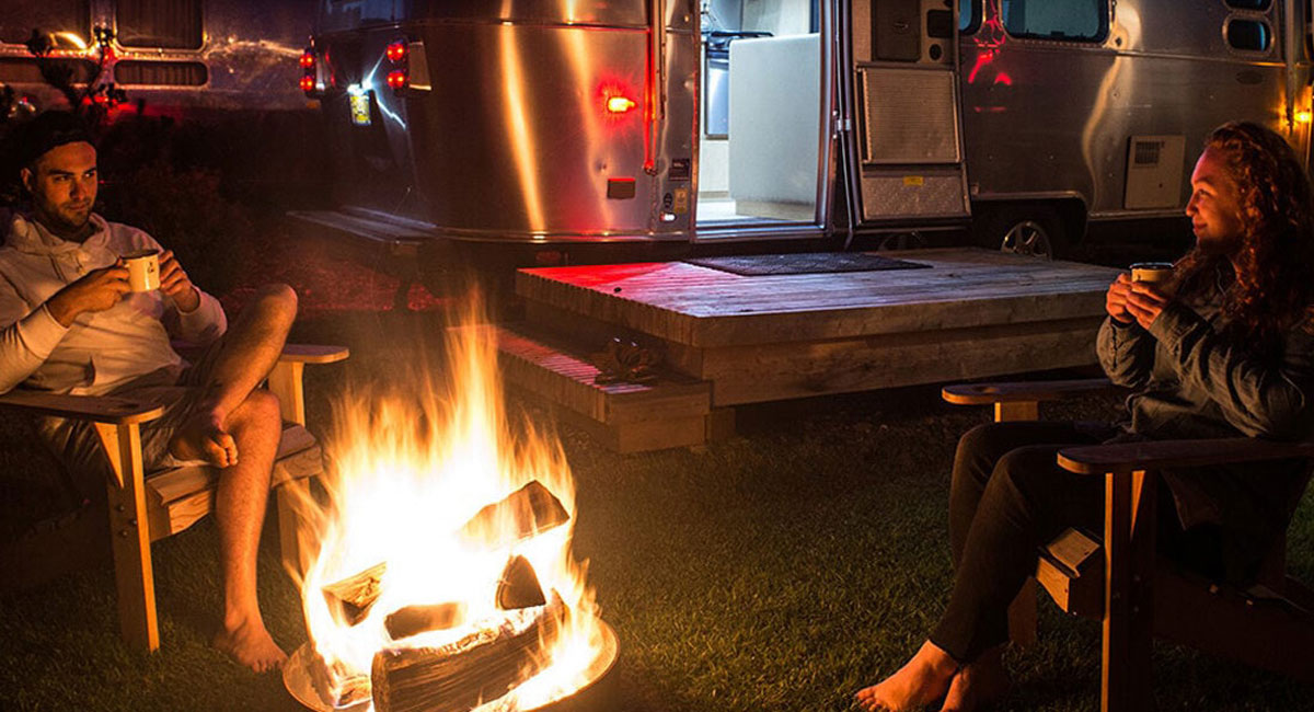A couple sits around a campfire in front of an airstream trailer.