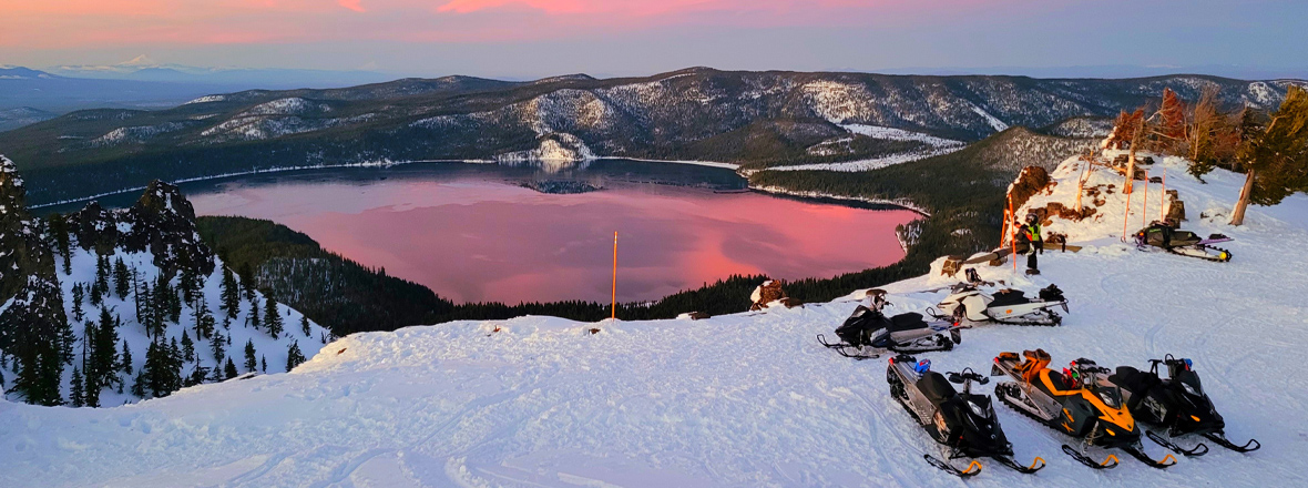 Snowmobiles parked on a snowy mountain overlooking a lake reflecting a pink sunset, surrounded by forested hills.