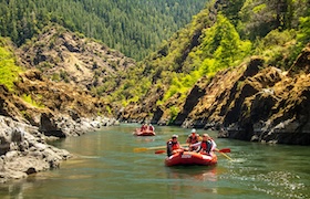 Groups of people rafting in red inflatable boats through a scenic river canyon surrounded by lush green forest.