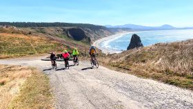 Four cyclists riding on a coastal road with sweeping views of cliffs and the ocean.