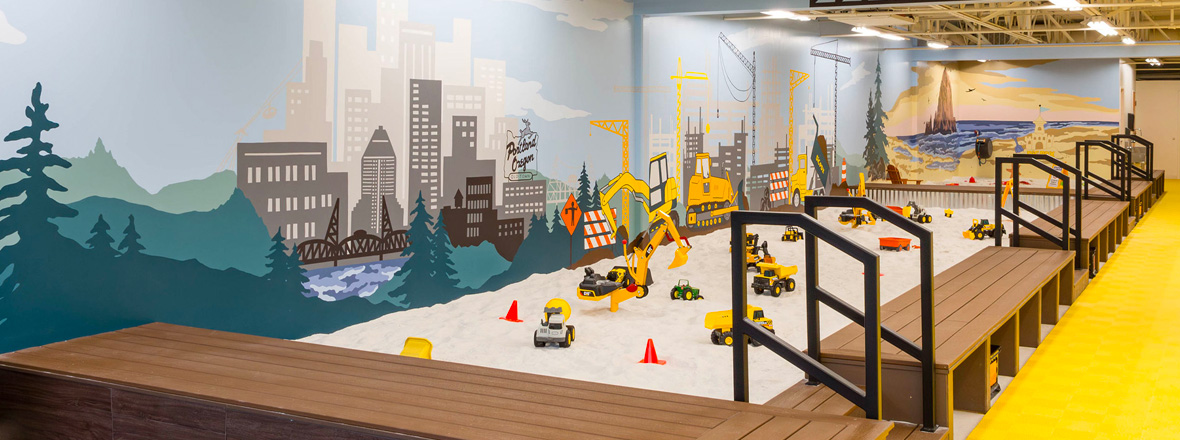 An indoor sandpit with toy construction trucks. The wall is a mural of Portland's skyline and more construction truck imagery.
