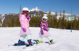 Two young children dressed in colorful ski outfits and tutus, skiing on a snowy slope.