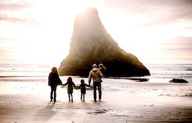 A family walking hand-in-hand on a sandy beach at sunset, facing a towering sea stack with sunlight streaming through.
