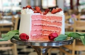 A layered pink cake with white frosting, topped with strawberries, displayed on a silver cake stand