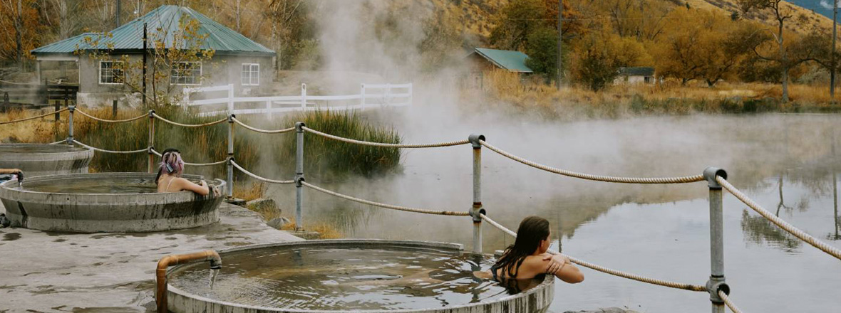 Women relaxing in circular soaking tubs outdoors set against a view of a natural hot springs.