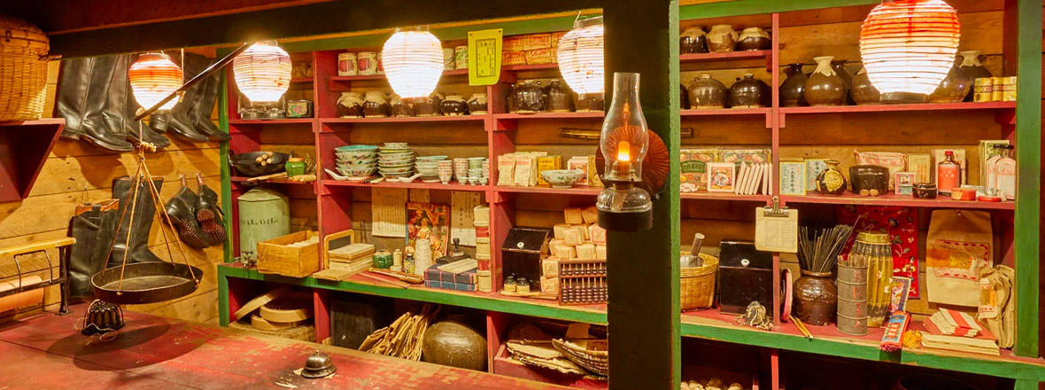 A historic general store display with wooden shelves stocked with pottery, packaged goods, and household items, illuminated by hanging paper lanterns and an oil lamp.