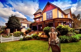 The Gilbert House Children's Museum, a vibrant Victorian-style house with a white picket fence and a bronze statue of a child sitting on a bench in the foreground.