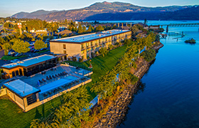 An aerial view of the Hood River Inn, located along the Columbia River, featuring a pool, green lawns, and a bridge with mountains in the background.