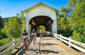 Cyclists riding across a historic white covered bridge surrounded by lush greenery under a bright blue sky.