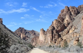 A rugged desert canyon landscape with towering rock formations, a dirt road and a bright blue sky