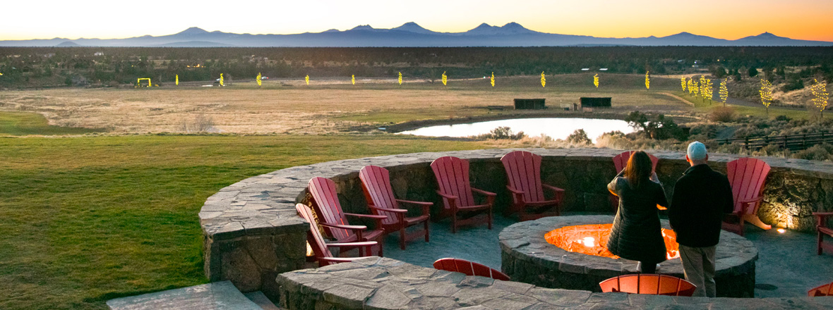 A couple stands next to an outdoor fireplace with mountains in the distance.