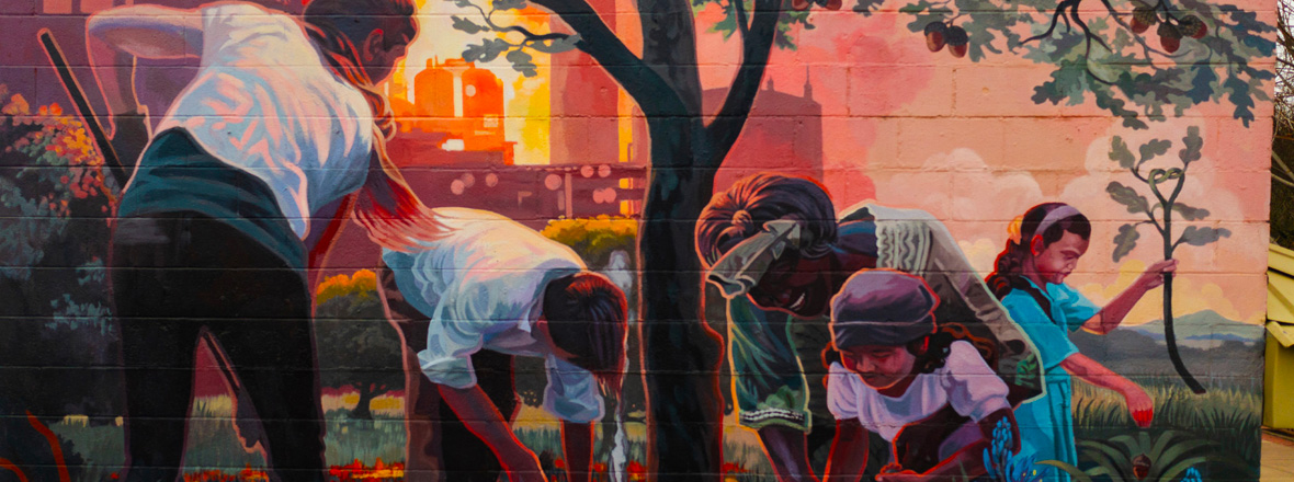 A colorful mural depicting people of different ages working together in a garden, with a tree in the foreground and an industrial cityscape in the background.