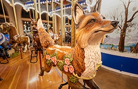 A detailed fox figure decorated with floral designs on a wooden carousel, surrounded by other animal figures and bright lights.