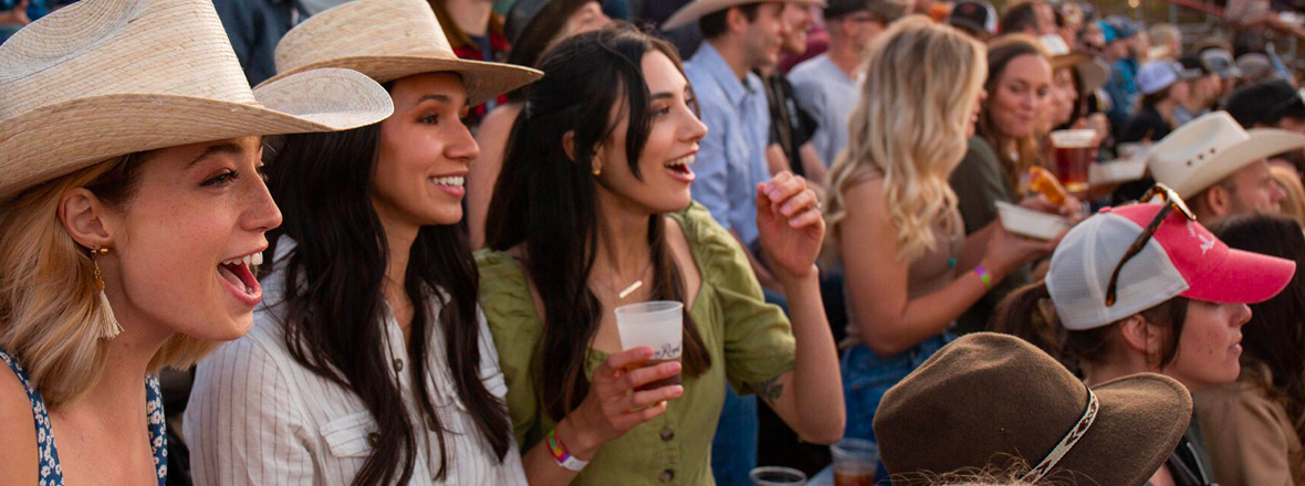A lively crowd, with women in cowboy hats holding drinks, cheers and enjoys an outdoor event.