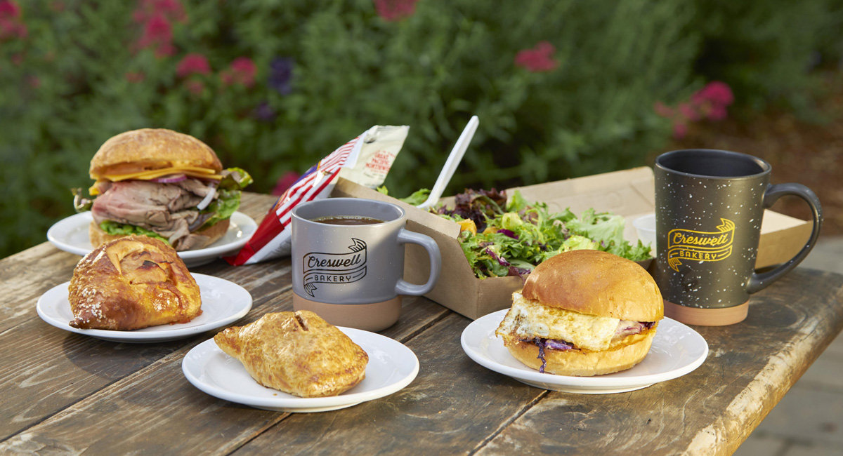 Several pastries, sandwiches and mugs emblazoned with Creswell Bakery on a table.