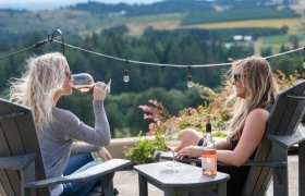 Two women enjoying glasses of rosé on a patio overlooking rolling vineyard hills, with string lights adding a relaxed ambiance.