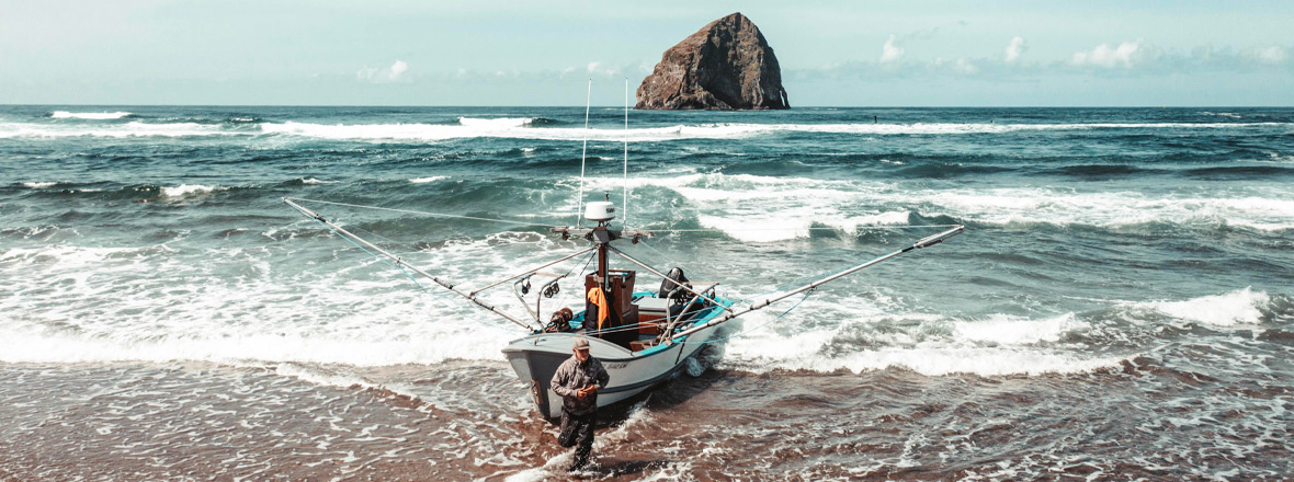 A man in front a a beached fishing boat. In the background, the ocean surrounds a large sea stack.