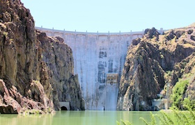Tall concrete dam set between rugged canyon walls, with calm green water in the foreground under a clear sky.